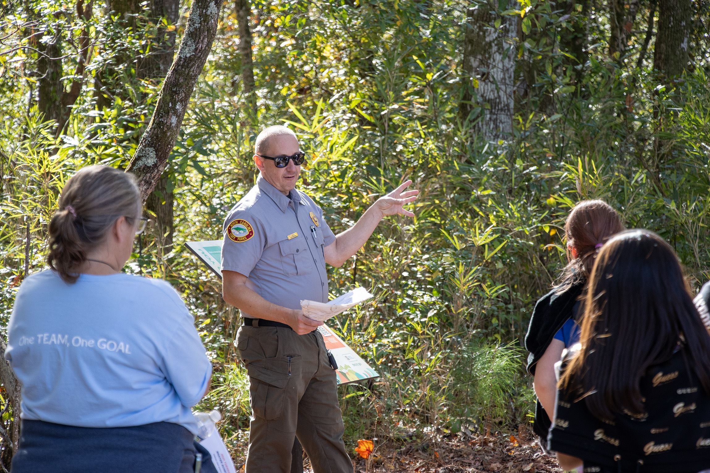 An NC Park Ranger, standing in front of a wooded area, facing a group of students and teaching them about wetlands. He is using hand movements for emphasis